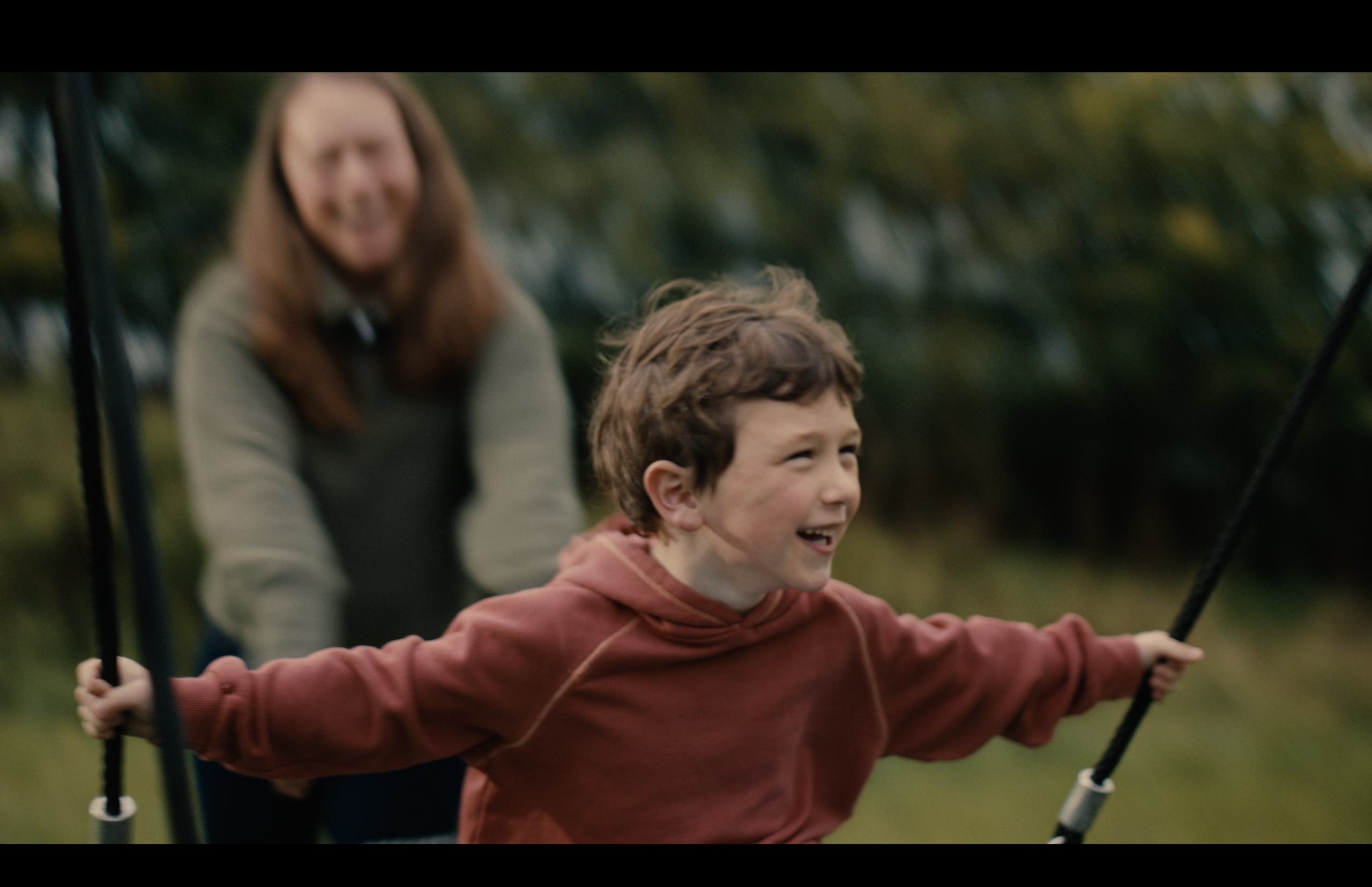 Still from CHAS TV advert featuring the playpark at Robin House hospice. A boy in a rust-coloured hoodie on an outdoor swing smiles with delight. Behind him is an adult woman with long hair who is blurred but you can still see the smile on her face too.