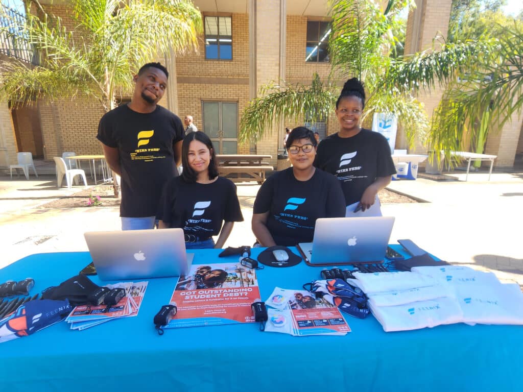 "Nix fees" - four students at a Feenix table outdoors wear black tshirts with the 'Nix fees' campaign message. 
