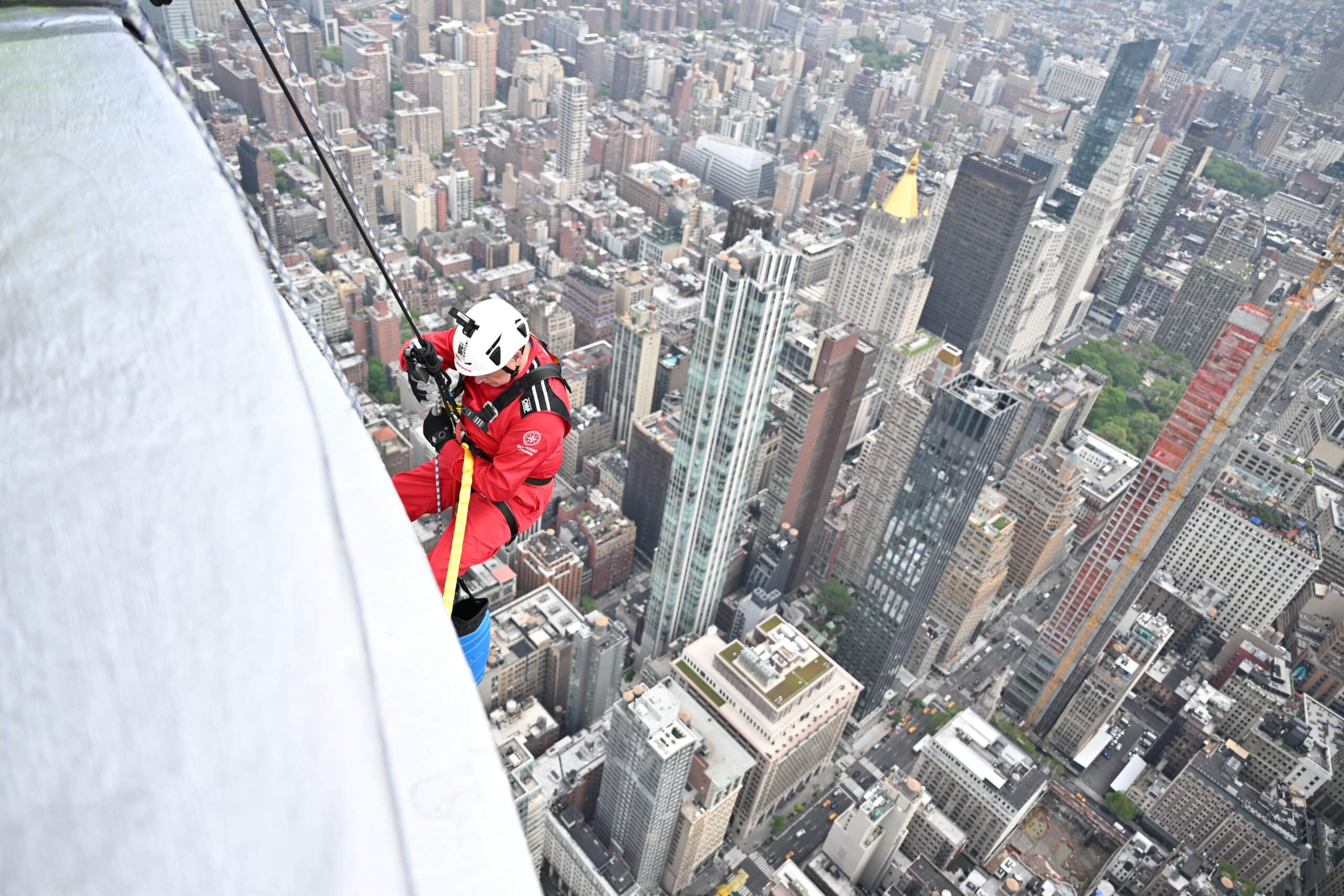 An abseiler in red suit steps off at the top of the Empire State Building, with New York City far below.