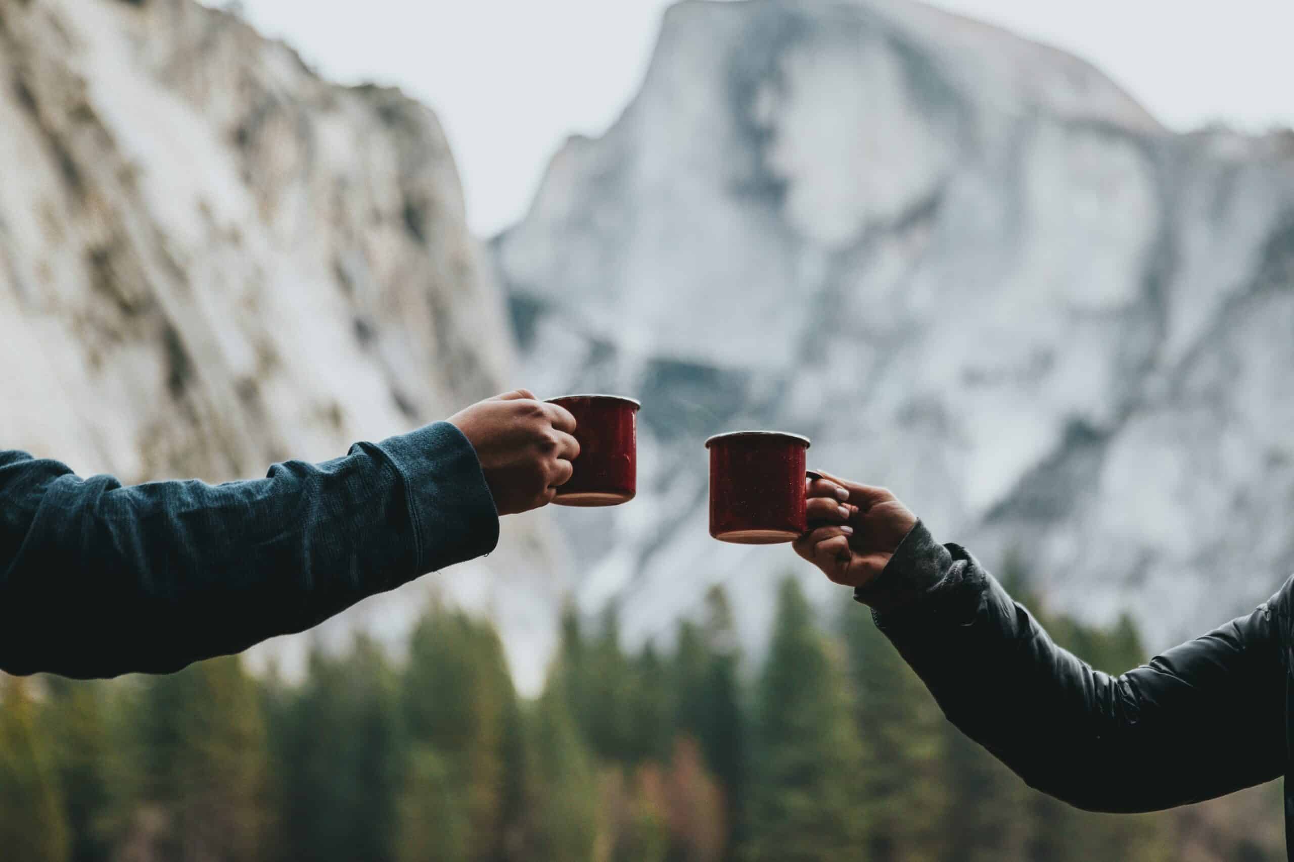 Two people clink cups of coffee, with just their arms showing, against a backdrop of mountains and for trees.